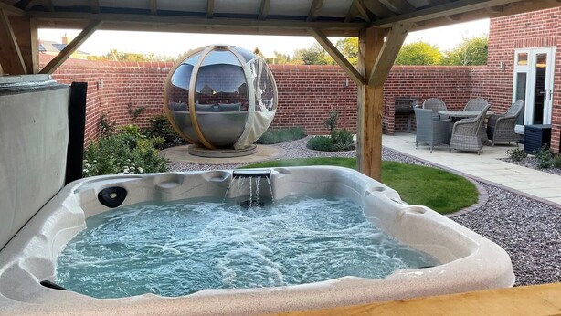 Een hottub onder een houten pergola, een bolvormige spiegel en een bakstenen muur op de achtergrond.