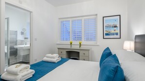 Een hotelkamer met een bed, een nachtkastje, een raam met zonwering, een lijst met een foto aan de muur, en een badkamer zichtbaar door een glazen deur.