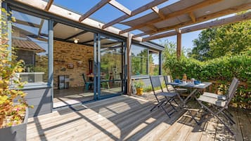 Una terraza de madera con mobiliario exterior y un ventanal de vidrio que da a una habitación con paredes de piedra y una mesa.