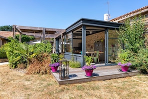 A covered outdoor area with a wooden deck, potted plants, and a view of the interior.