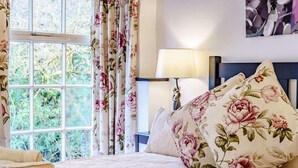 Une chambre à coucher avec un lit, des rideaux à motifs floraux, une lampe de chevet et une vue sur la verdure à l’extérieur.