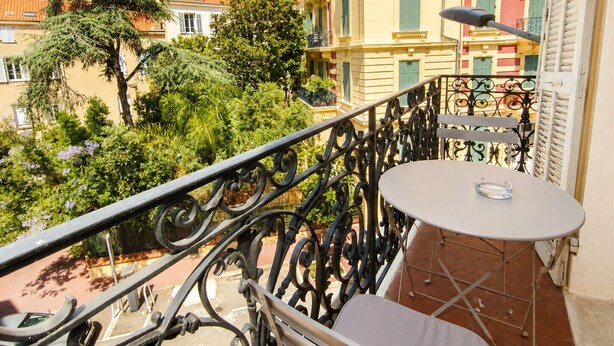 Un balcone con un tavolo rotondo e una sedia, affacciato su un cortile alberato con edifici.