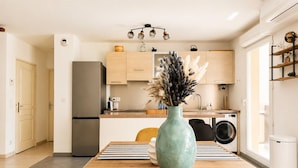 A modern kitchen with wooden cabinets, a stainless steel refrigerator, a washing machine, and a vase with dried flowers on the dining table. - Sunny Terrace & Open Views | Pool, Parking, A/C | Cannes – BENAKEY (Cannes)