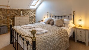 Ein Schlafzimmer mit gewölbter Decke, einer Steinwand, einem Bett mit gesteppter Decke, Nachttischen mit Lampen und einem Dachfenster.