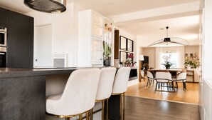 Een moderne keuken met een centraal eiland, barkrukken, een eethoek met tafel en stoelen, en een groot raam.