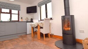 A modern kitchen with grey cabinets, a dining table with chairs, a TV mounted on the wall, and a wood-burning stove. - 1 Bed in Launceston (oc-79379) (Davidstow)