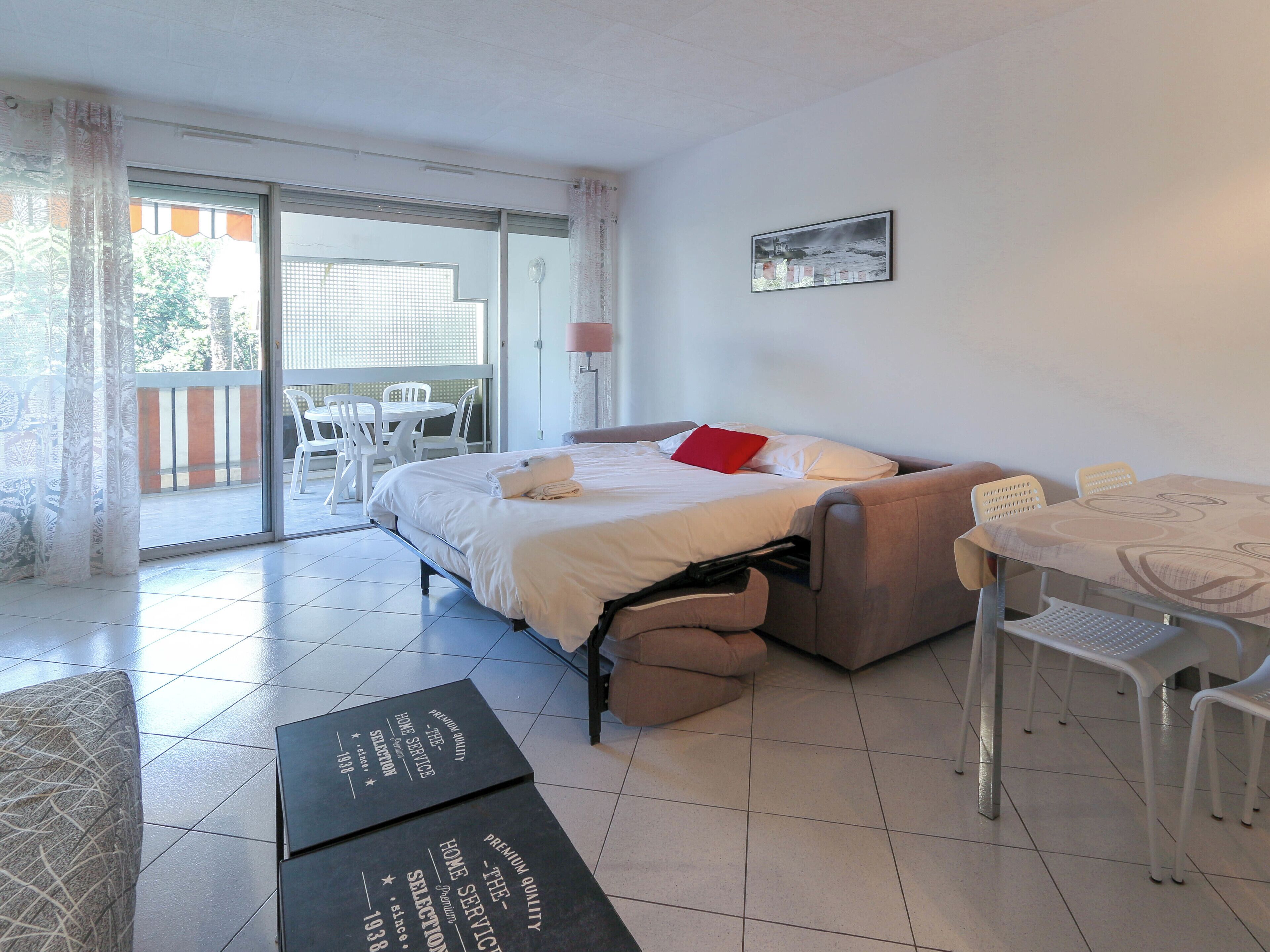 A hotel room with a bed, a sofa, a dining table with chairs, a balcony with a table and chairs, and a wall-mounted lamp.