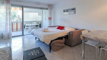 A hotel room with a bed, a sofa, a dining table with chairs, a balcony with a table and chairs, and a wall-mounted lamp.