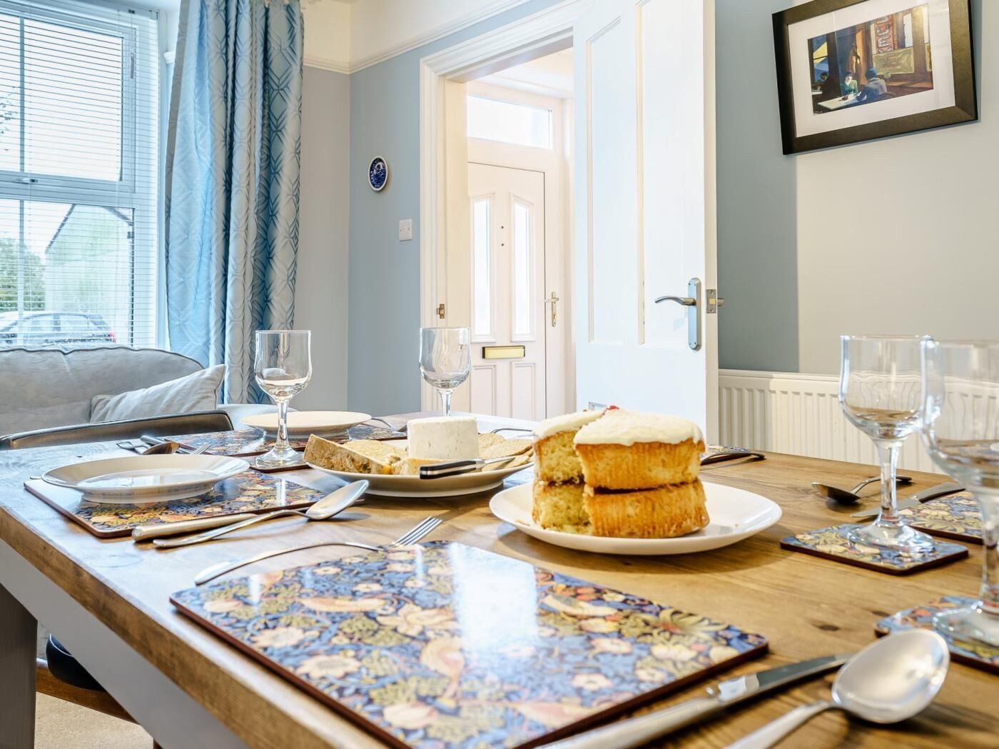A dining table set with plates, glasses, and cutlery, with a cake and a window in the background.