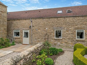 A stone building with a tiled roof, two windows, a door, and a small garden area with plants and a gravel path. - 2 Bed in Scarborough (oc-79523) (Sawdon)