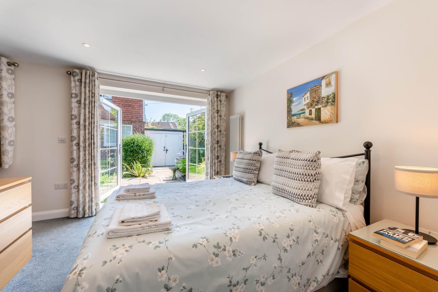 A bedroom with a bed, a bedside table with a lamp, a painting on the wall, and a view of the garden through the door.