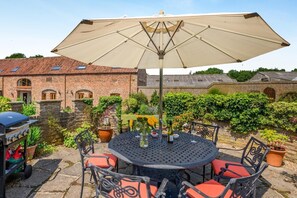 Een terras met een ronde tafel en stoelen, een barbecue en een grote parasol.