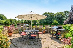 Een terras buiten met een ronde tafel, stoelen, een grote parasol en een barbecue.