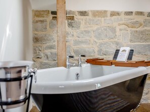 Une salle de bain avec un mur en pierre, une baignoire blanche et un plateau en bois sur lequel repose un livre.