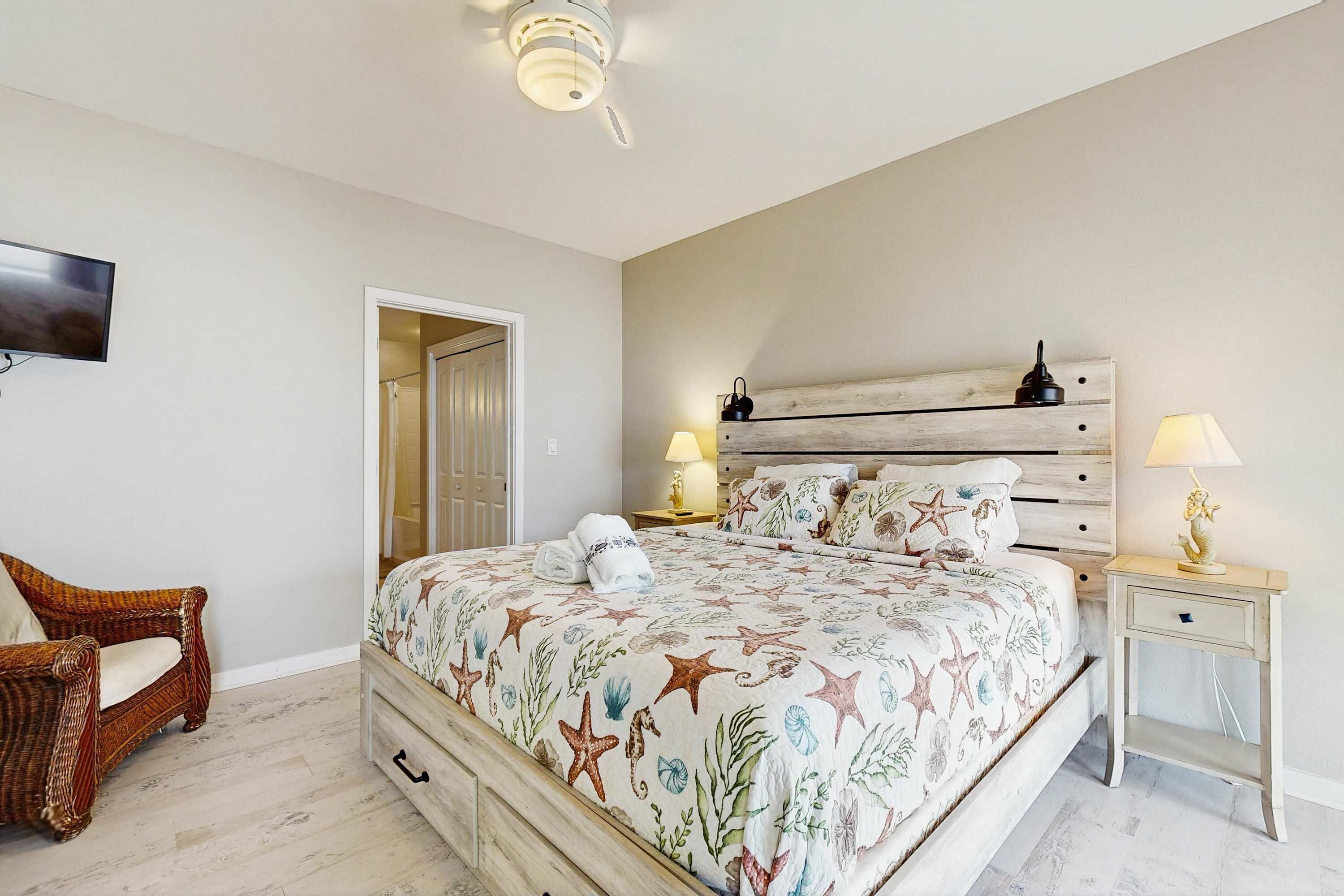 Un dormitorio con una cama, dos mesitas de noche, un televisor, una silla de mimbre y una puerta que da a otra habitación.
