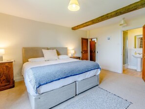 Une chambre à coucher avec un lit, des tables de chevet, une poutre en bois au plafond et une salle de bain visible à travers une porte ouverte.