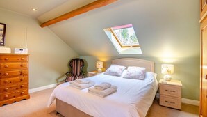 Un dormitorio con techo inclinado, una cama con almohadas, una silla, un aparador, lámparas de noche y una ventana.