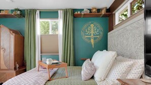 Um quarto com uma cama, cômoda de madeira, mesa de cabeceira com bandeja, janela com cortinas e um painel decorativo na parede.