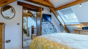 Ein Schlafzimmer mit schrägen Wänden, einer Steinwand, einem Bett mit floralem Überwurf, einer hölzernen Kommode und einem Dachfenster.