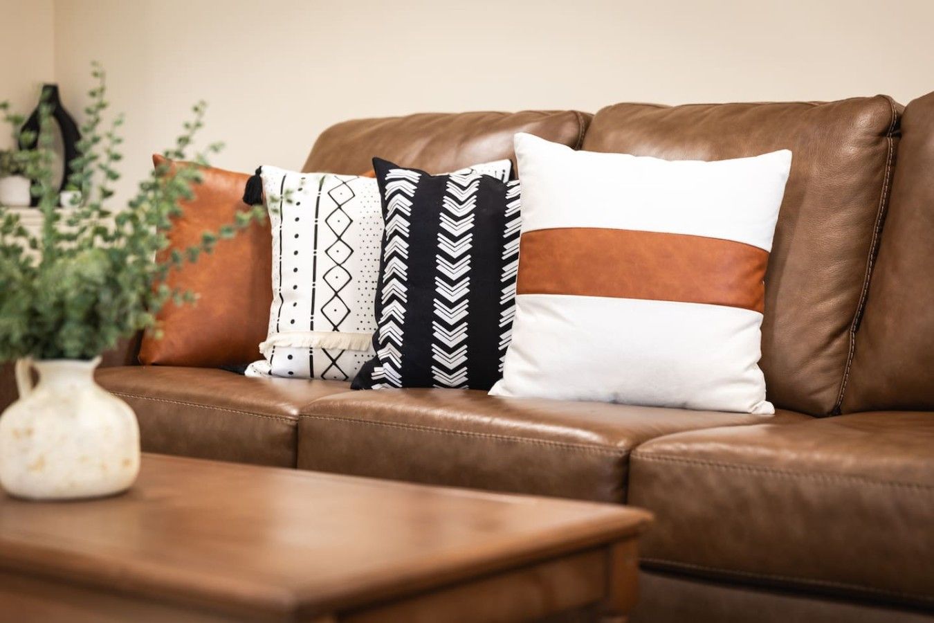 A brown leather sofa with patterned cushions and a white vase with green plants on a wooden table.