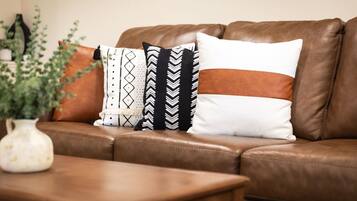 A brown leather sofa with patterned cushions and a white vase with green plants on a wooden table.