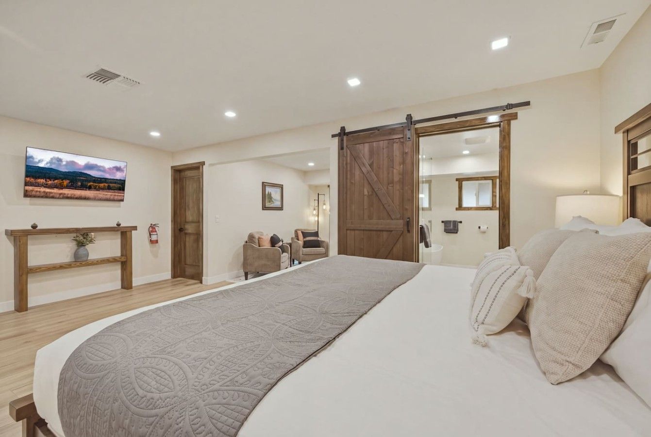 Een slaapkamer met een groot bed, een houten vloer, een televisie aan de muur, een schuifdeur in boerderijstijl en een klein zithoekje met stoelen.