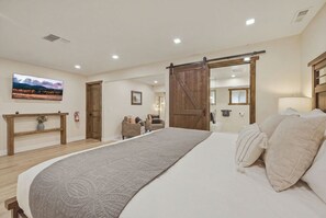 Un dormitorio con una cama grande, piso de madera, un televisor en la pared, una puerta corrediza estilo granero y una pequeña zona de estar con sillas.