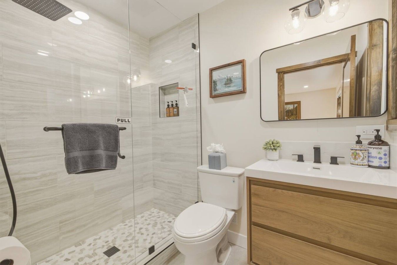 A modern bathroom with a glass-enclosed shower, a toilet, a sink with toiletries, and a mirror.