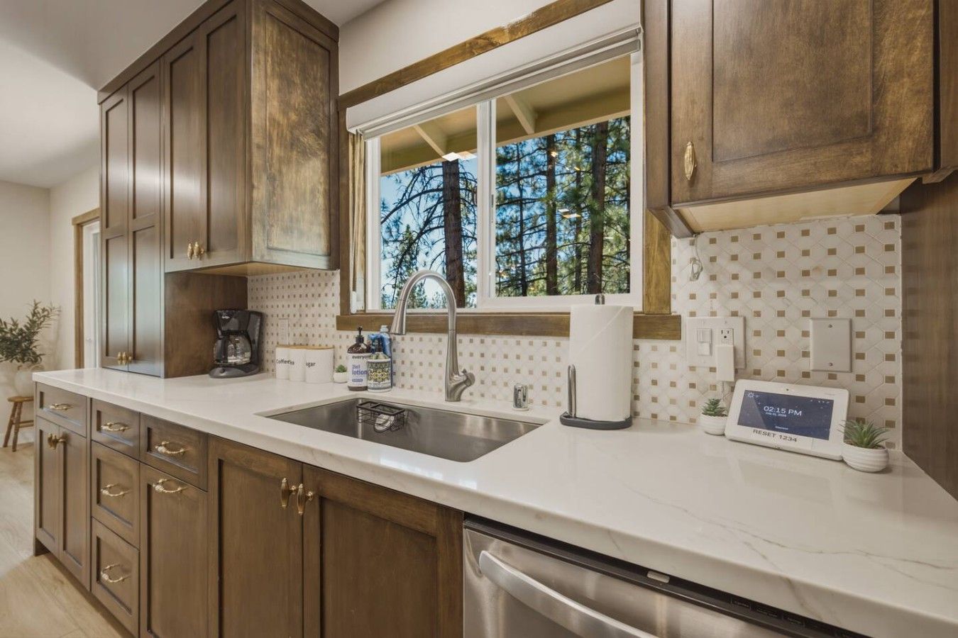 Una cocina con armarios de madera, un lavamanos de acero inoxidable, una ventana con vistas a árboles y un reloj digital que muestra la hora.