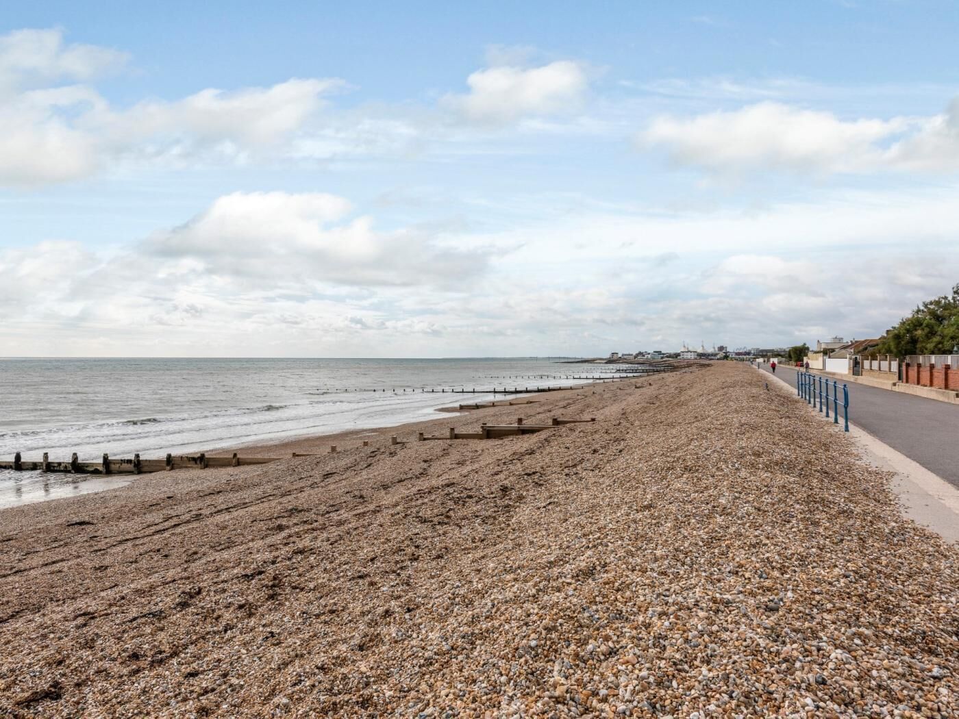 Ein Kiesstrand mit blauem Himmel, eine parallel zum Strand verlaufende Straße und Gebäude in der Ferne.