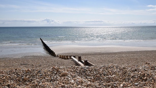Ein Kiesstrand mit einem hölzernen Steg, der sich ins Meer erstreckt, unter einem wolkenlosen Himmel.