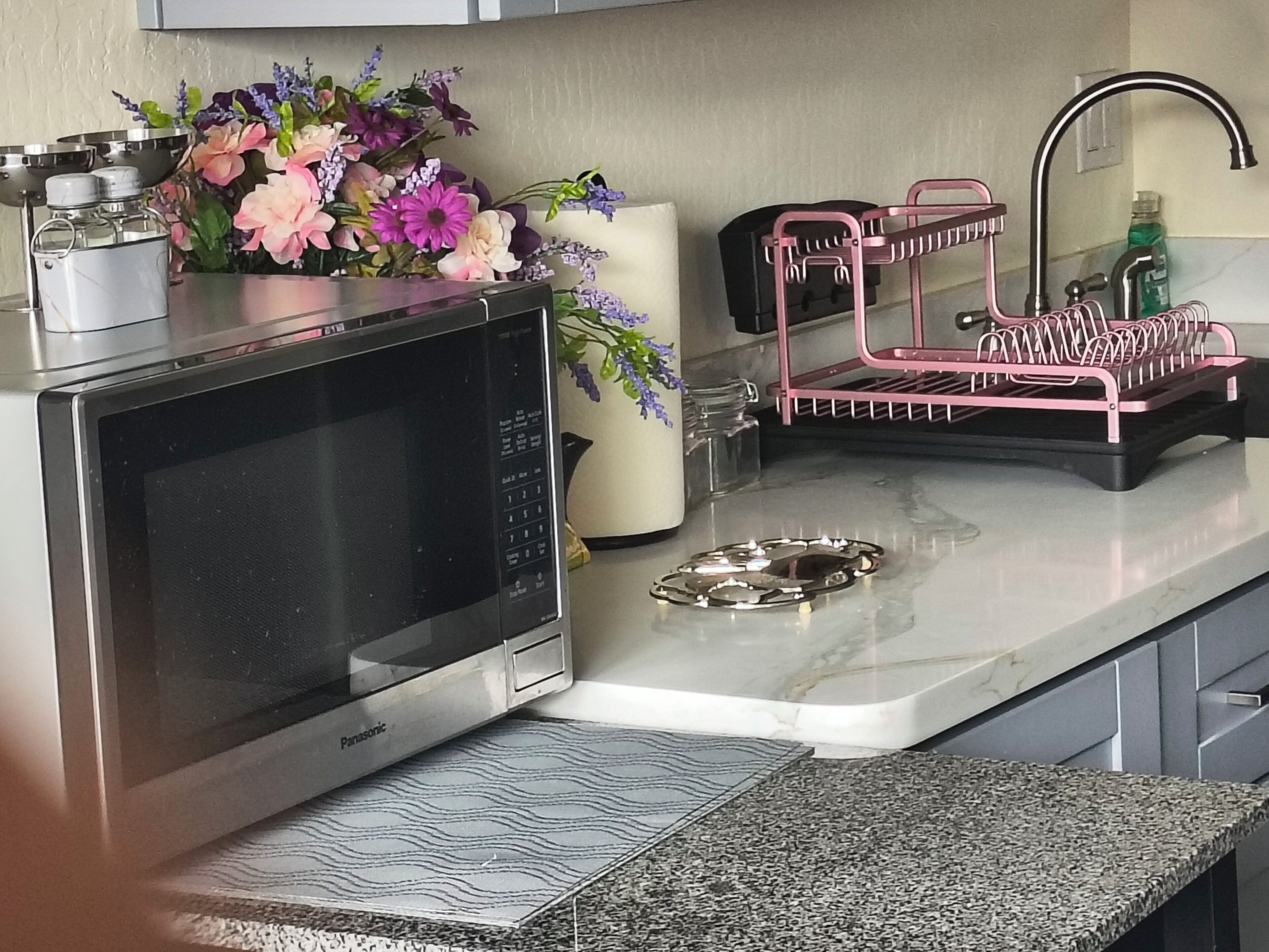 A kitchen with a microwave, a pink utensil holder, a vase with flowers, and a bottle of dish soap.