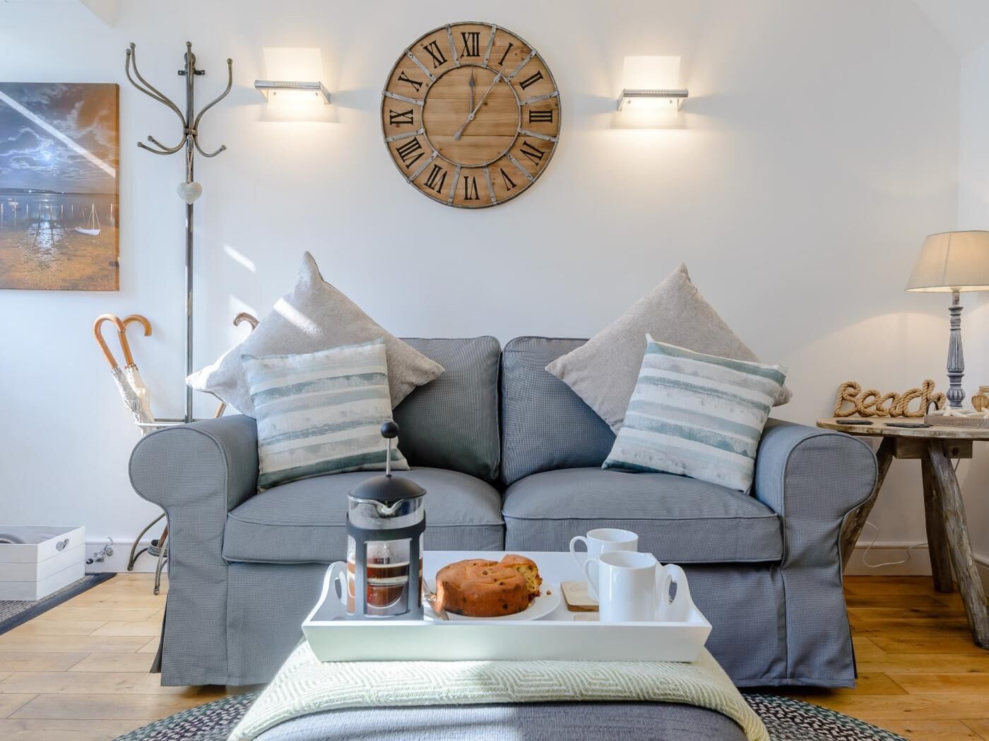 A living room with a grey sofa, striped pillows, a wooden clock, and a tray with a muffin and cups.