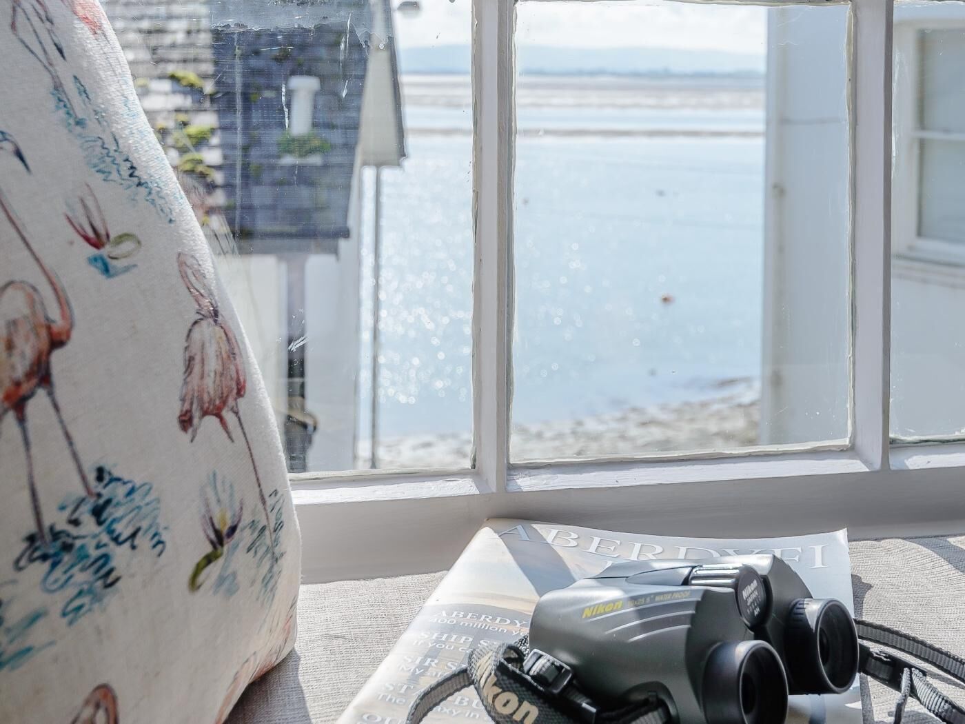 A window with a view of the sea, a Nikon camera on a surface, and a patterned fabric.