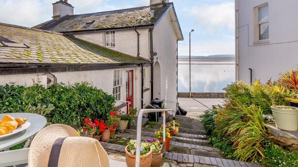 Une rue étroite avec un bâtiment blanc, une porte rouge et une vue sur la mer. On y trouve des plantes en pot et une table avec des croissants.