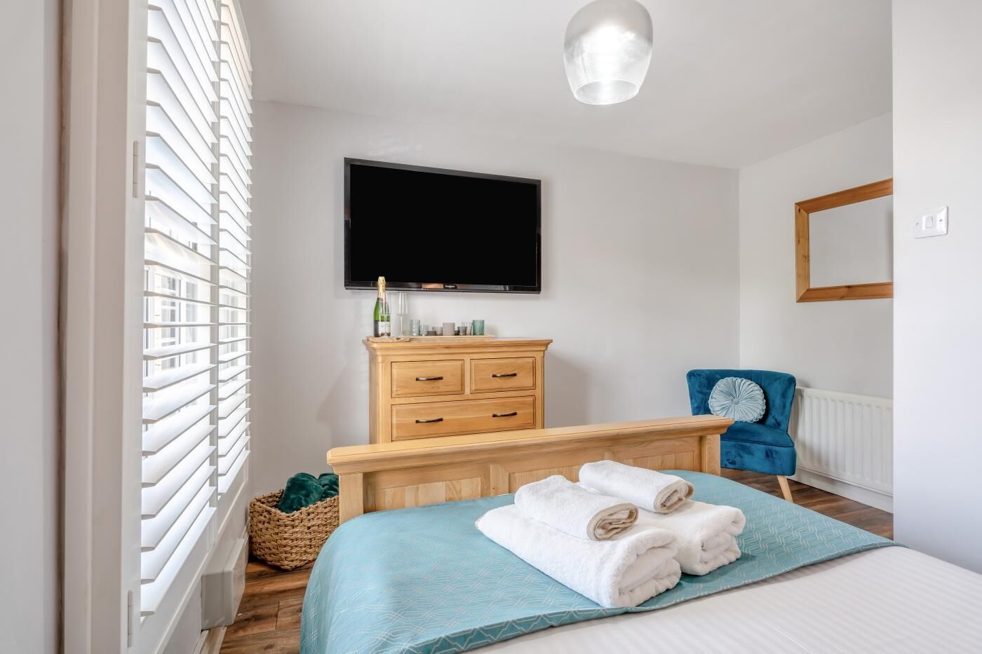 A bedroom with a bed, a wooden dresser, a television, a blue chair, and a window with blinds.