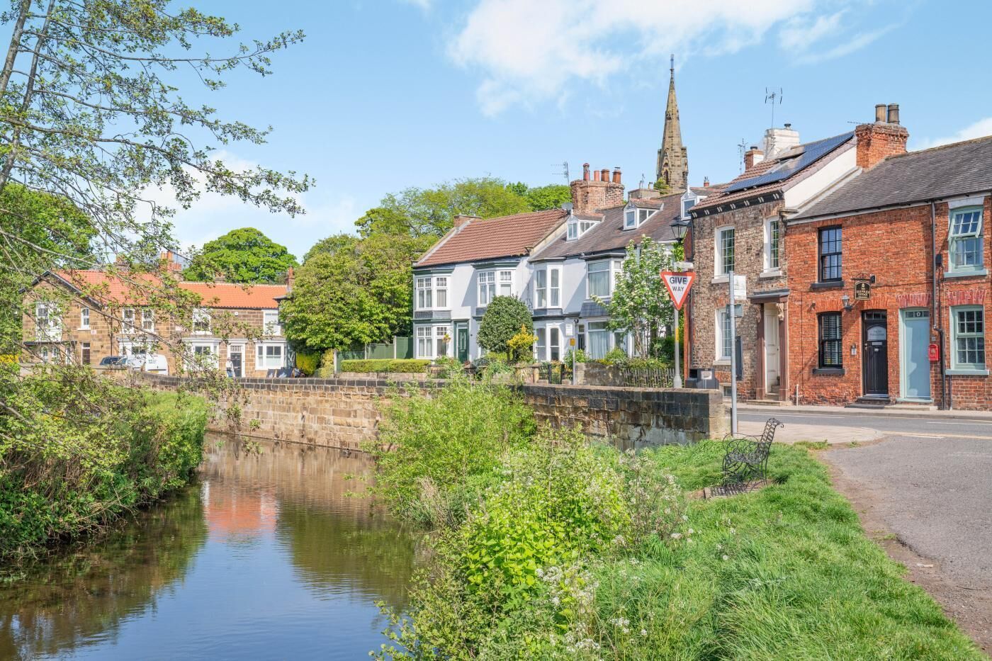 A riverside town with historic buildings, a church spire, and a 'Give Way' sign.