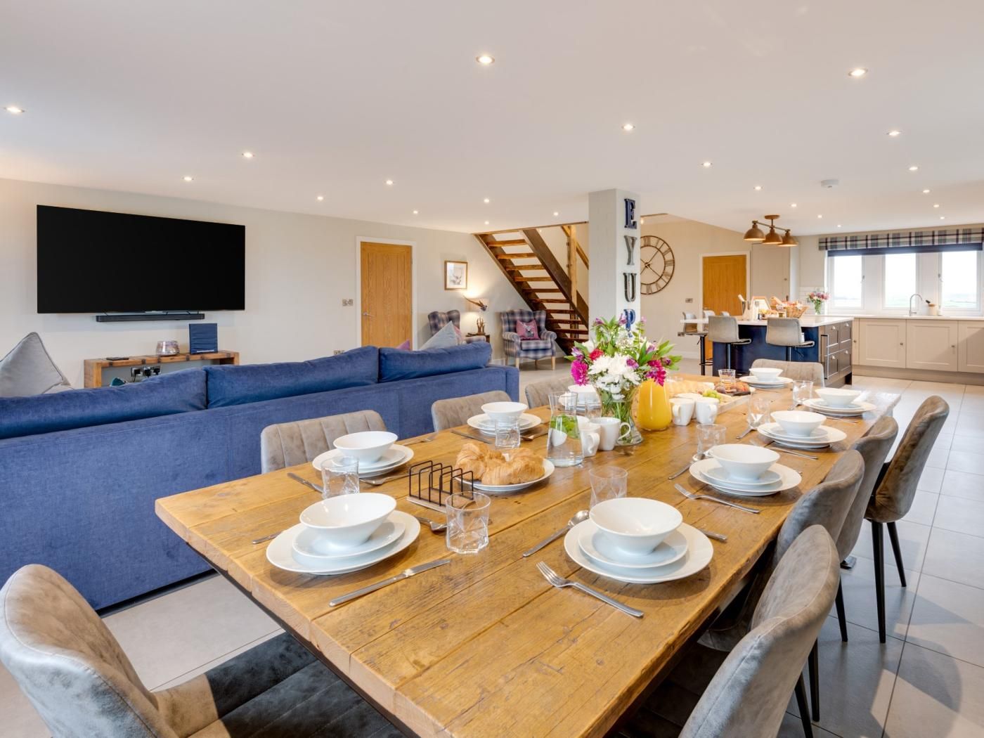 A modern living room with a large sofa, a dining table set for four, and a kitchen in the background.