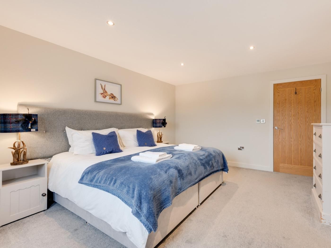 Une chambre à coucher comprenant un lit, des tables de chevet, une commode, une porte en bois et un luminaire fixé au mur.