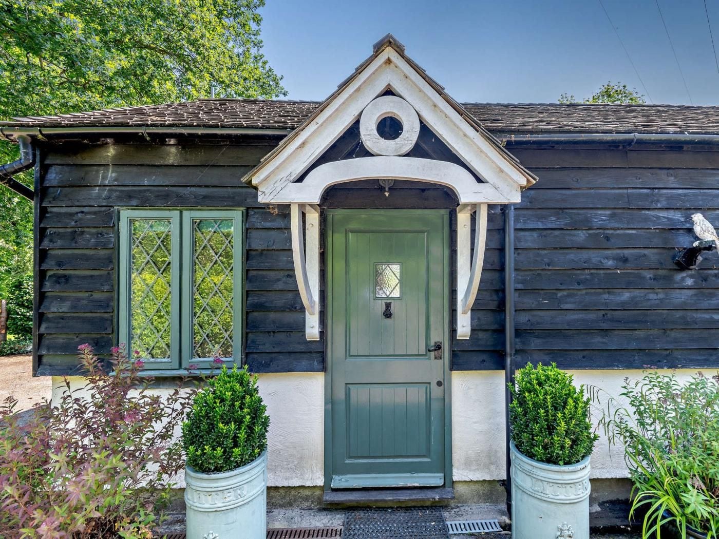 A wooden house with a green door, two windows with shutters, and a small porch area with potted plants.