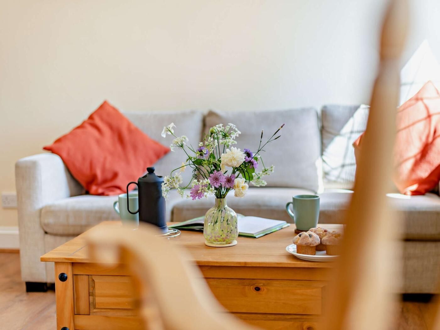 Ein gemütliches Wohnzimmer mit einem Holztisch, einer Blumenvase, einer Kaffeekanne, einer Tasse und einem Tablett mit Muffins.