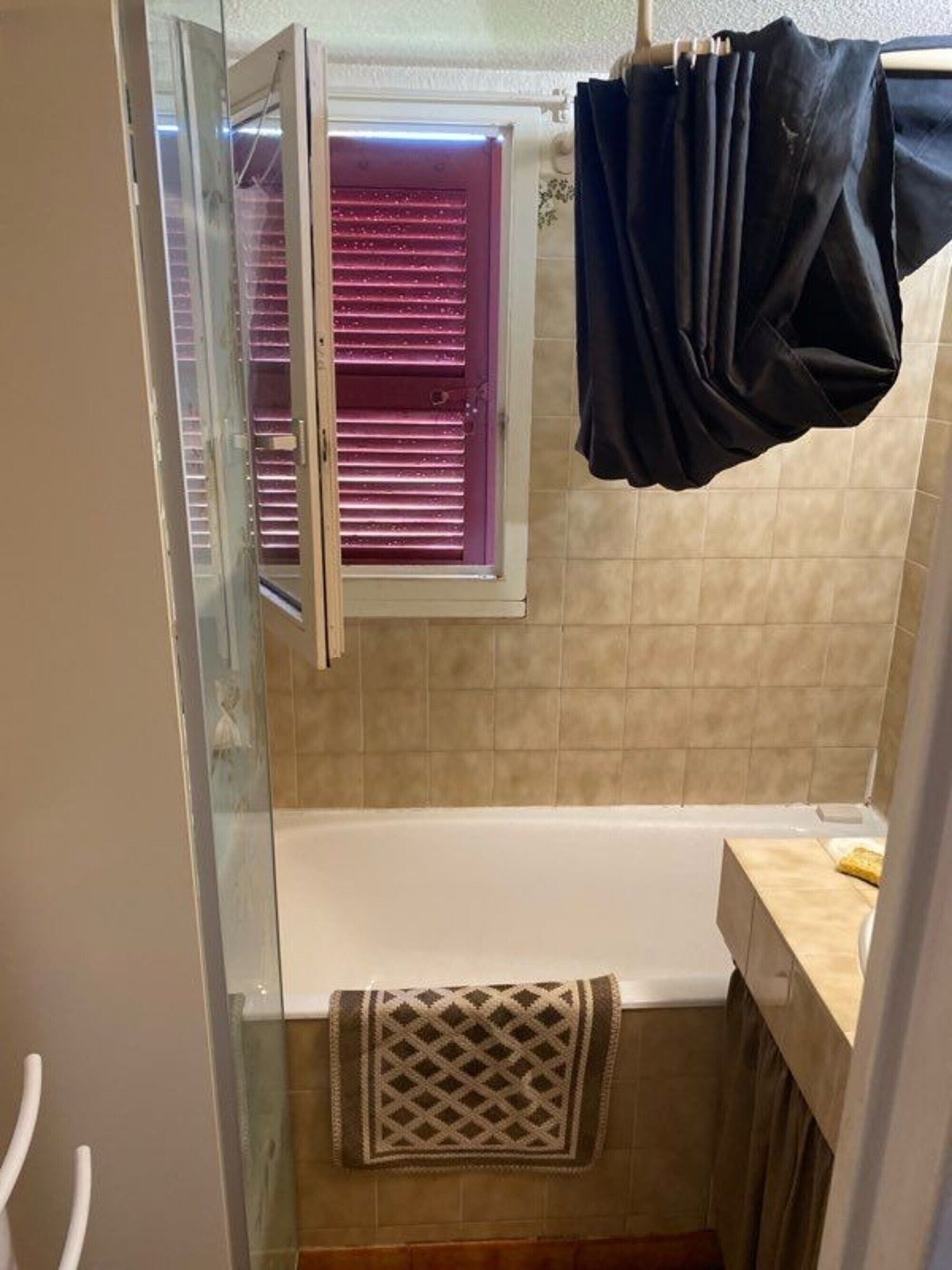 Une salle de bain avec une baignoire, un espace de douche en verre, un porte-serviettes et une fenêtre avec des volets.