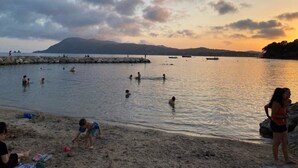 Eine Strandansicht mit badenden und entspannten Menschen, einem steinernen Wellenbrecher und einem Berg im Hintergrund.