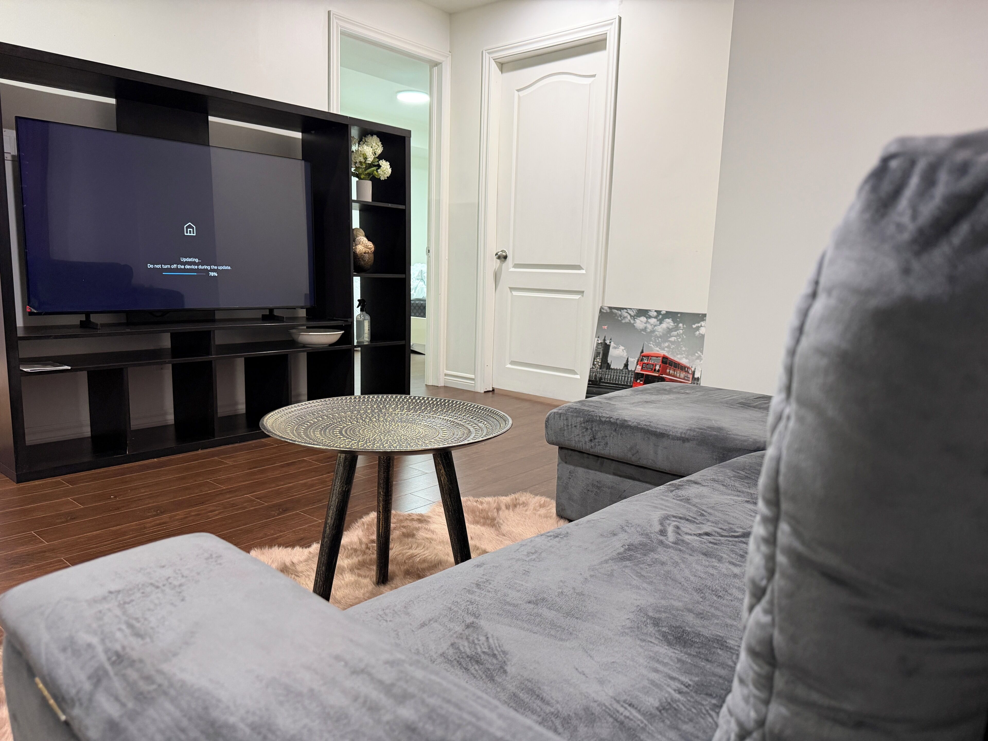 Een woonkamer met een flatscreen televisie, grijze banken, een kleine ronde tafel en een witte deur.