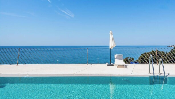 Ein klares, blaues Schwimmbecken mit Blick aufs Meer, ein weißer Sonnenschirm und ein Metallzaun.