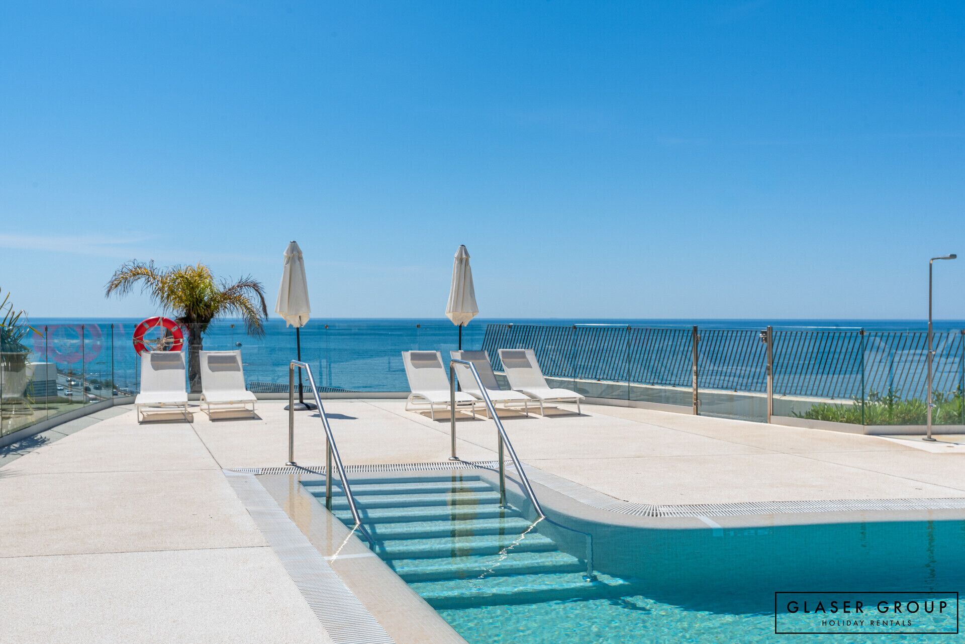 A poolside area with lounge chairs and umbrellas, a clear view of the ocean, and a glass barrier.
