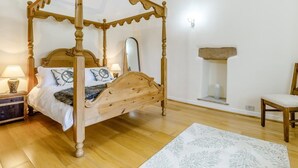 Ein Bett mit Baldachin, ein hölzernes Kopfende, ein Nachttisch mit Lampe, ein Sessel und ein Schlafplatz für Haustiere an der Wand.