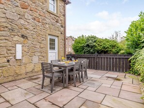 Ein Steinhaus mit Tür und Fenster, eine Terrasse mit Tisch und Stühlen sowie ein Holzzaun.
