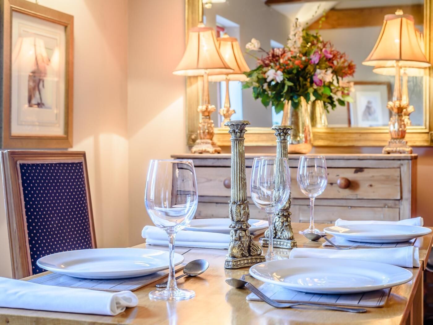 A dining table set with plates, glasses, and cutlery, with a mirror and floral centerpiece in the background.