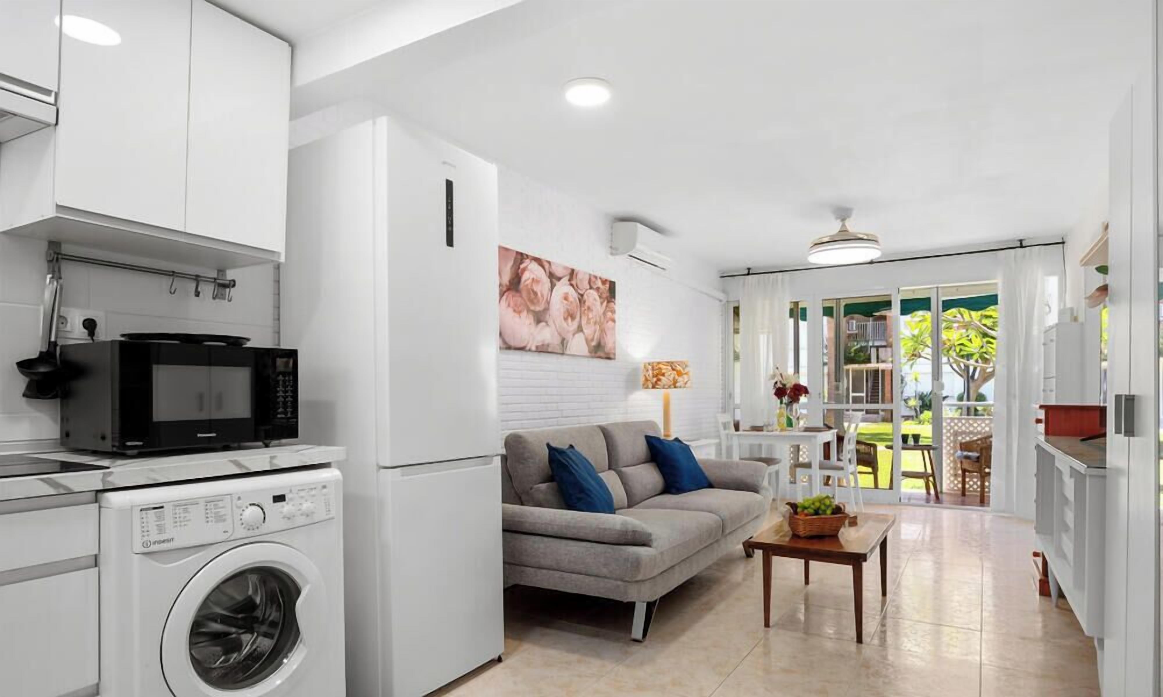 A compact living space with a kitchenette, a washing machine, a grey sofa with blue cushions, and a dining area with a table and chairs.